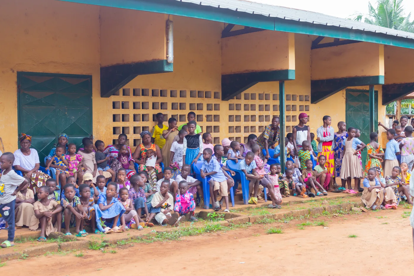 Gruppenfoto von Helfern, lokalen Vertretern und vielen Kindern mit Schulranzen und Schulmaterial, die ein Banner mit der Aufschrift „Kossivi's HOPE" halten, vor Schulgebäuden und einer bewaldeten Bergkulisse.