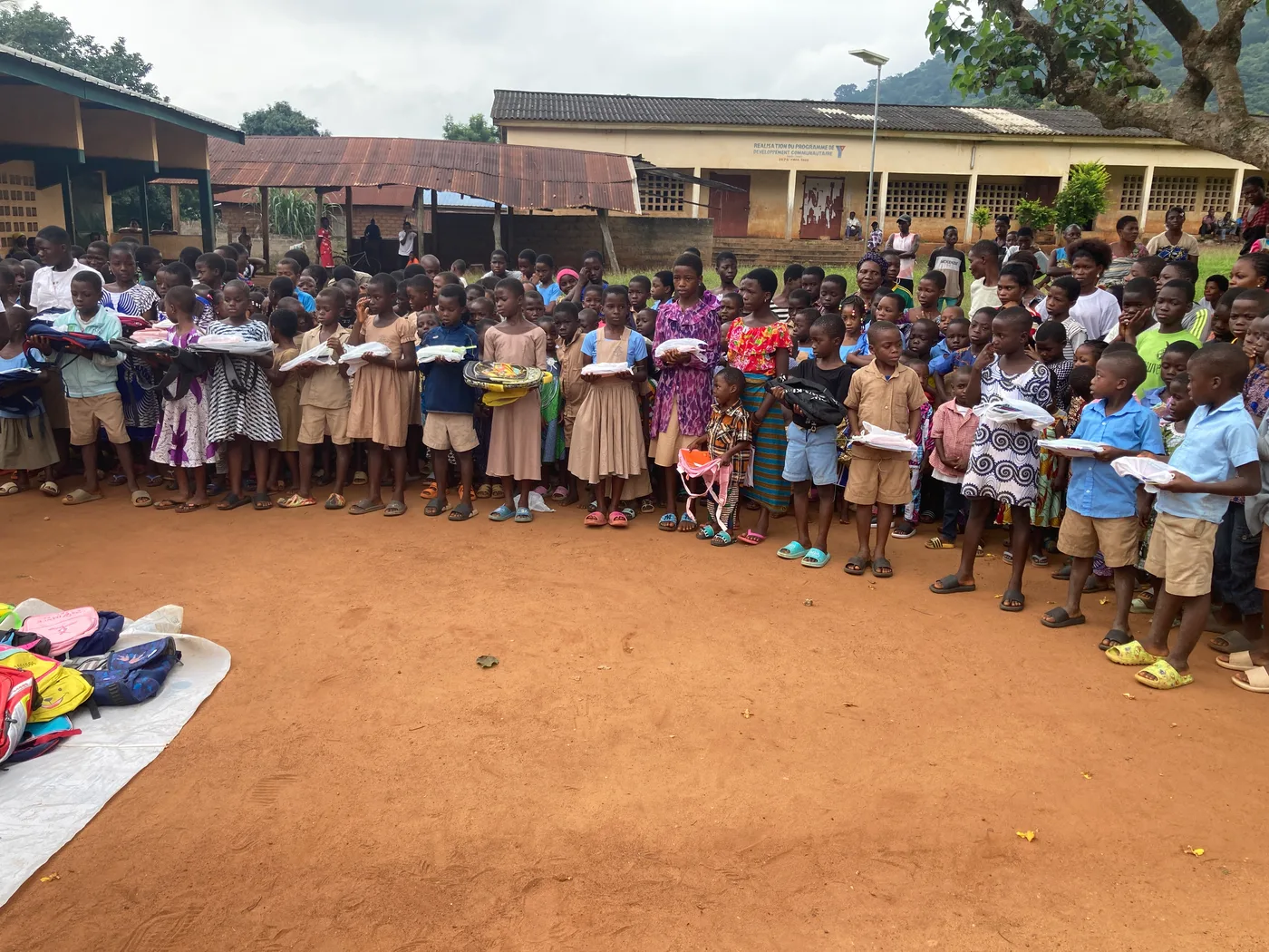 Gruppenfoto von Helfern, lokalen Vertretern und vielen Kindern mit Schulranzen und Schulmaterial, die ein Banner mit der Aufschrift „Kossivi's HOPE" halten, vor Schulgebäuden und einer bewaldeten Bergkulisse.