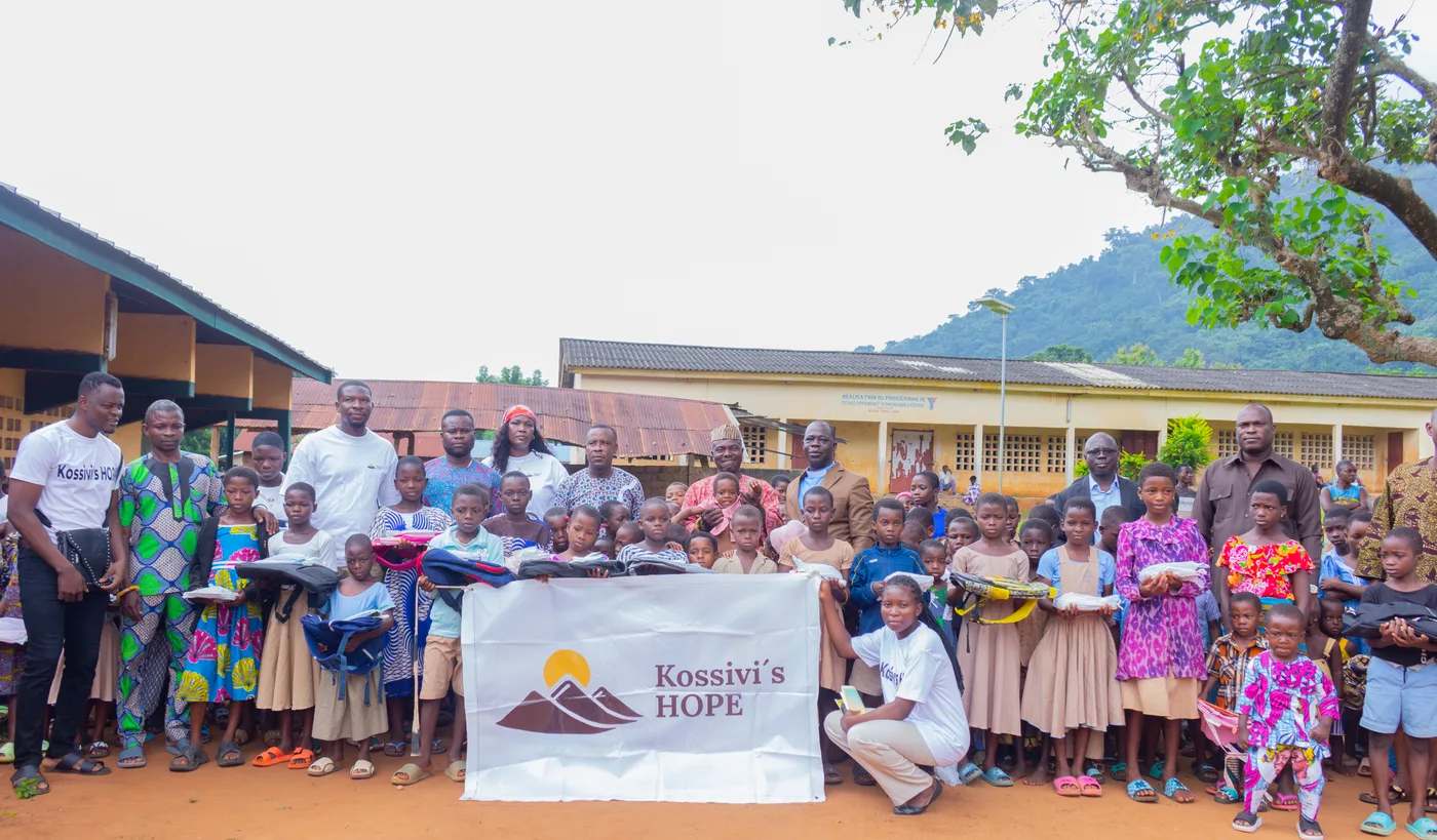 Gruppenfoto von Helfern, lokalen Vertretern und vielen Kindern mit Schulranzen und Schulmaterial, die ein Banner mit der Aufschrift „Kossivi's HOPE" halten, vor Schulgebäuden und einer bewaldeten Bergkulisse.